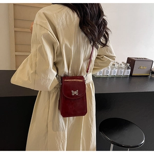 Detail of wine red leather crossbody phone bag with silver butterfly charm on beige coat