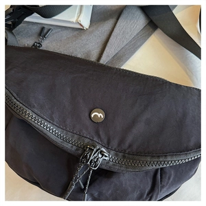 Detail of a minimalist black nylon crossbody bag with dual zippers and a circular logo