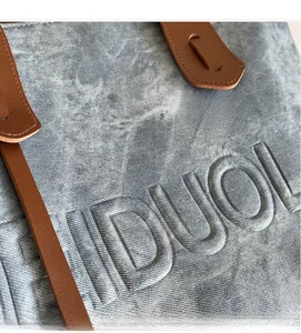 Detailed close-up of washed blue denim tote bag with embossed IDUOL logo and brown leather strap