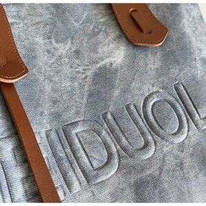 Detail of stone-washed denim tote bag featuring 3D embossed IDUOL lettering and brown leather strap