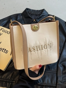 Fashionable beige tote bag with brown trim, featuring letter embroidery and a gold twist lock