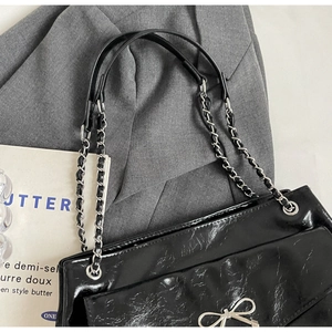 Detail of black glossy leather tote featuring silver bow and chain strap on a blazer