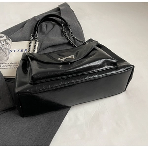 Detail of black glossy tote bag featuring silver bow clasp and chain strap on grey fabric