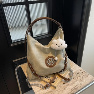 Detail of canvas tote bag with brown leather trim, metal M logo, and plush cloud charm