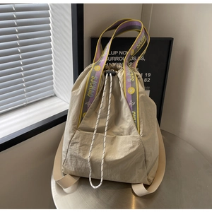 Detalle de mochila beige claro con cordón y correas de logo moradas y amarillas en mesa