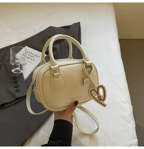 Close-up of cream bowling bag with heart charm on white bedding and grey blazer