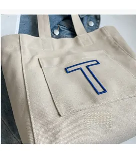 Close-up of cream canvas tote bag with blue embroidered letter T and front pocket detail
