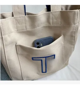 Close-up of beige canvas tote bag front pocket with blue T embroidery and smartphone