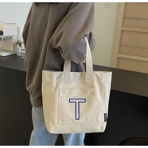 Detail of cream canvas tote bag featuring large blue embroidered letter T and brand label