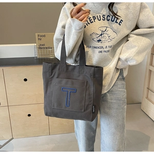 Detail of dark gray canvas tote bag featuring blue embroidered letter T and brand tag