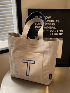 Minimalist beige canvas tote bag with blue embroidered T-letter pocket and contrast handles