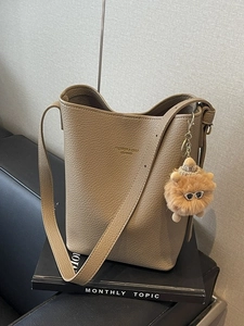 Beige litchi leather bucket bag with plush charm and long strap in a modern studio setting