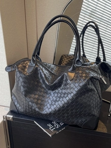 Large black woven tote bag with dual leather handles and reinforced base on a minimalist shelf