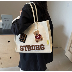 Detail of canvas tote bag showing plush bear patch embroidery and large capacity on shoulder