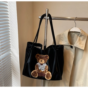 Detail of a black satin tote bag featuring a textured brown bear patch with striped shirt