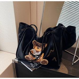 Detail of glossy black tote bag featuring a teddy bear plush patch and smooth mirror finish
