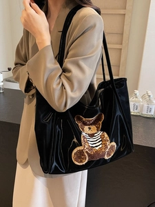 Black patent leather tote bag with large brown teddy bear patch and long shoulder straps
