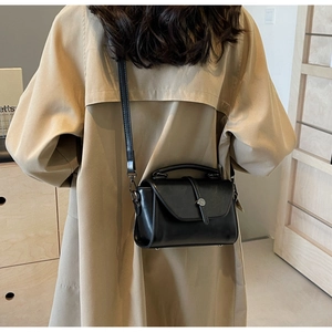 Detail of a black leather crossbody bag featuring a round metal lock and a beige trench coat