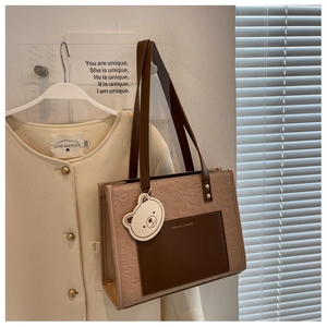 Detail der beigen Shopper-Tasche mit Bärenanhänger und Prägung vor warmem Hintergrund