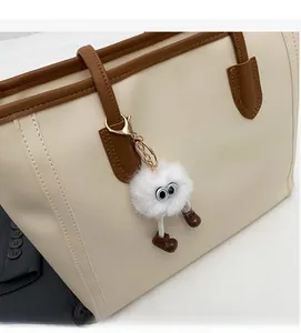 Detail of white plush cartoon charm with golden clasp on a beige leather tote bag