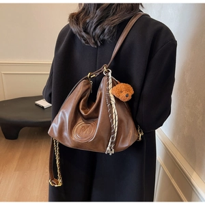 Detail of brown leather tote bag with M logo, gold chain, and cute teddy bear pendant