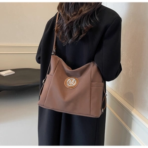 Detail of a brown tote bag with silver M logo badge and side pocket shown on a model