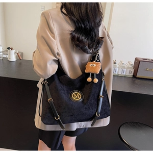 Detail of black corduroy tote bag with gold M logo, leather strap, and plush charm