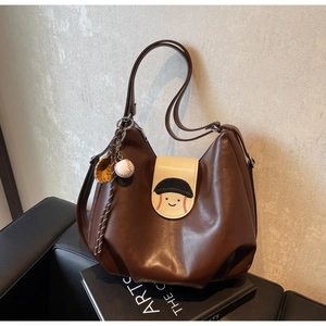 Detail of a brown leather tote bag with a baseball glove charm and cartoon face flap