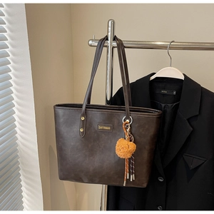 Detailaufnahme der braunen Tragetasche mit Metall-Logo und orangefarbenem Bommel