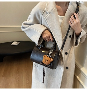 Detail of dark brown boston bag with golden plaque and teddy bear charm on beige coat