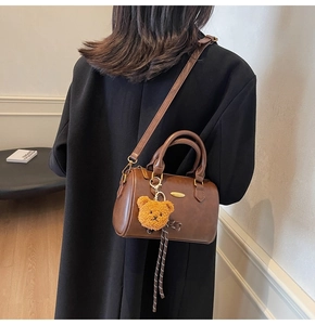 Detail of a brown Boston bag showing shoulder strap, teddy bear charm and metal plate