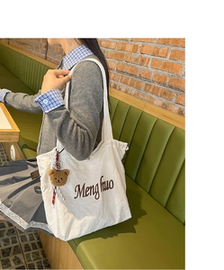 Close-up of a white canvas tote bag with Menghuo embroidery and a brown teddy bear charm