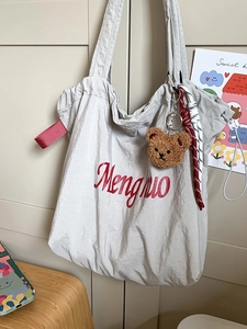 Minimalist grey tote bag with red embroidery and bear charm shown hanging on a white door