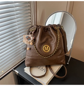 Close-up of brown leather bucket bag with gold M logo, chain detail and plush charm