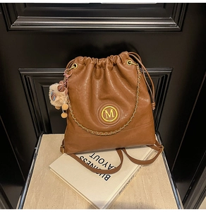 Close-up of brown bucket backpack with metallic M logo and plush charm on book