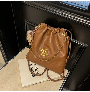 Detail of a brown leather drawstring backpack with gold M-logo, chain, and fur pom-pom