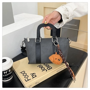 Gray trapezoid handbag with black handles and cute teddy bear charm on book background