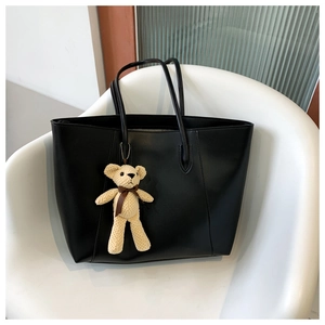 Close-up of a large black tote bag with a beige knitted bear charm on a white chair