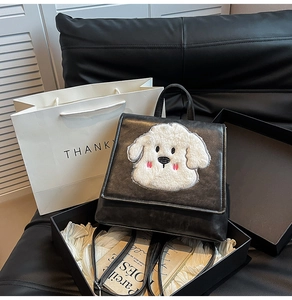 Detail view of a black faux leather backpack with a plush puppy patch in a gift box