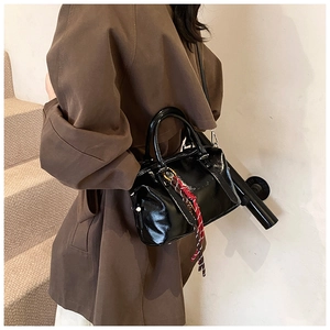Sac à main noir brillant avec pompon porté en bandoulière sur un trench-coat marron