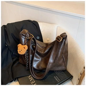 Detail of dark brown leather tote bag with wide strap and teddy bear charm on white chair