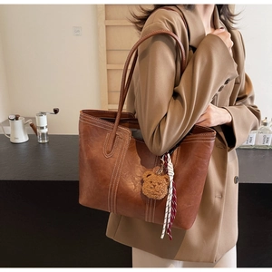 Detail of brown leather tote bag with white stitching and plush bear charm on shoulder