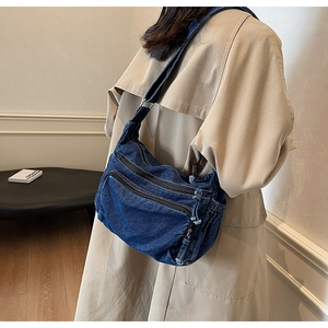 Detail of blue denim crossbody bag with multiple zippers worn over a khaki trench coat