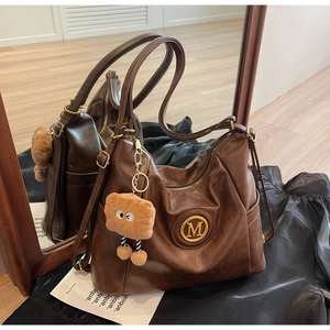 Detail of a brown leather tote bag featuring a gold M logo and funny plush toast pendant