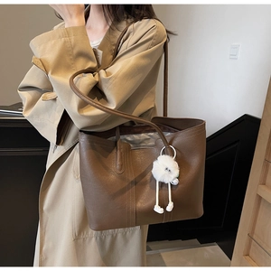 Brown leather tote bag with white plush charm shown on person wearing beige coat