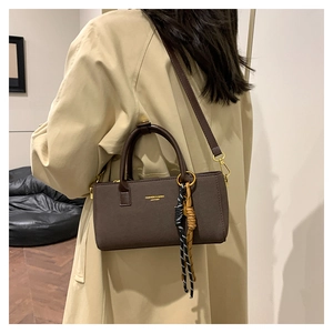 Deep brown crossbody bag with woven rope charms and gold lettering worn over a beige coat
