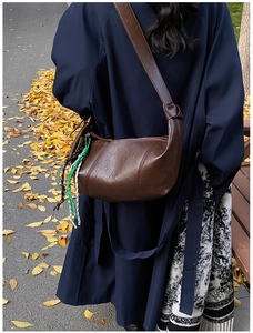 Brown leather crescent bag with woven charm shown on a shoulder in an autumn street setting