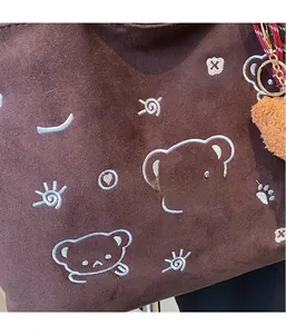 Close-up of brown suede tote bag with white cartoon embroidery and plush bear pendant