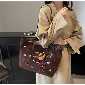 Close-up of brown suede tote bag featuring cartoon embroidery and plush ball pendant