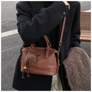 Close-up of a brown Boston bag with a golden metal logo and fuzzy pompom accessory